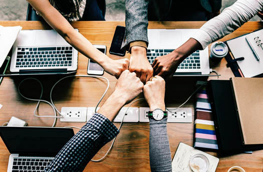 Five people sitting at a table full of laptops fist bumping each other